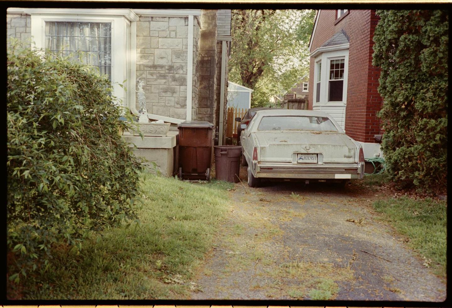 A white car is parked on a gravel driveway next to a house with a stone facade and a red brick house.