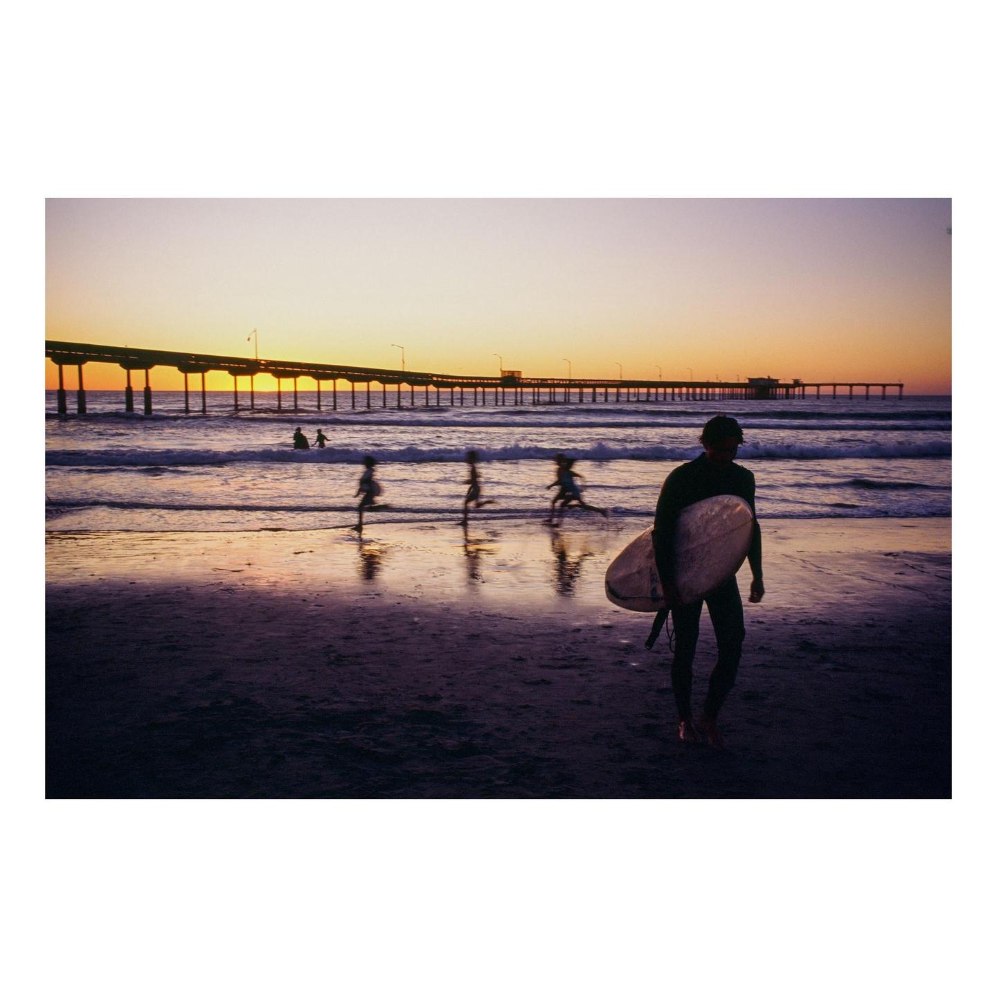 A surfer stands on a beach with a surfboard while children play in the water and a pier stretches into the distance at sunset.