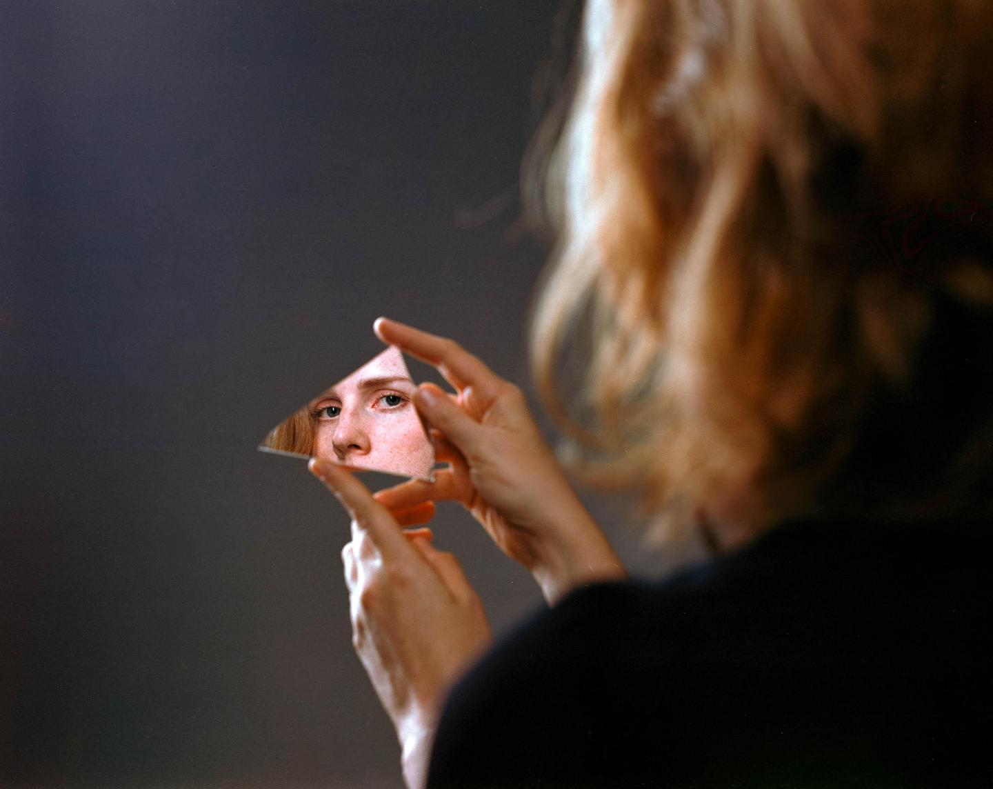 A woman holds a triangular piece of mirror reflecting her eye and nose.