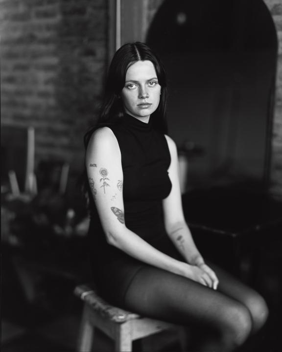 A woman with long hair and tattoos on her arms sits on a stool.