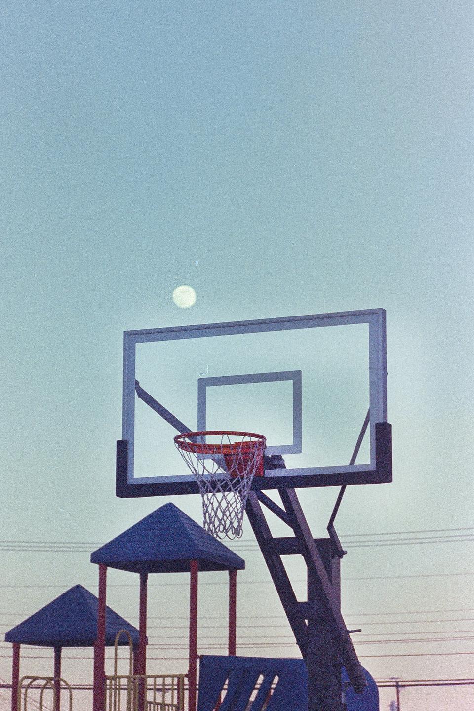 A basketball hoop and backboard are visible against a light blue sky with a full moon and playground equipment below.
