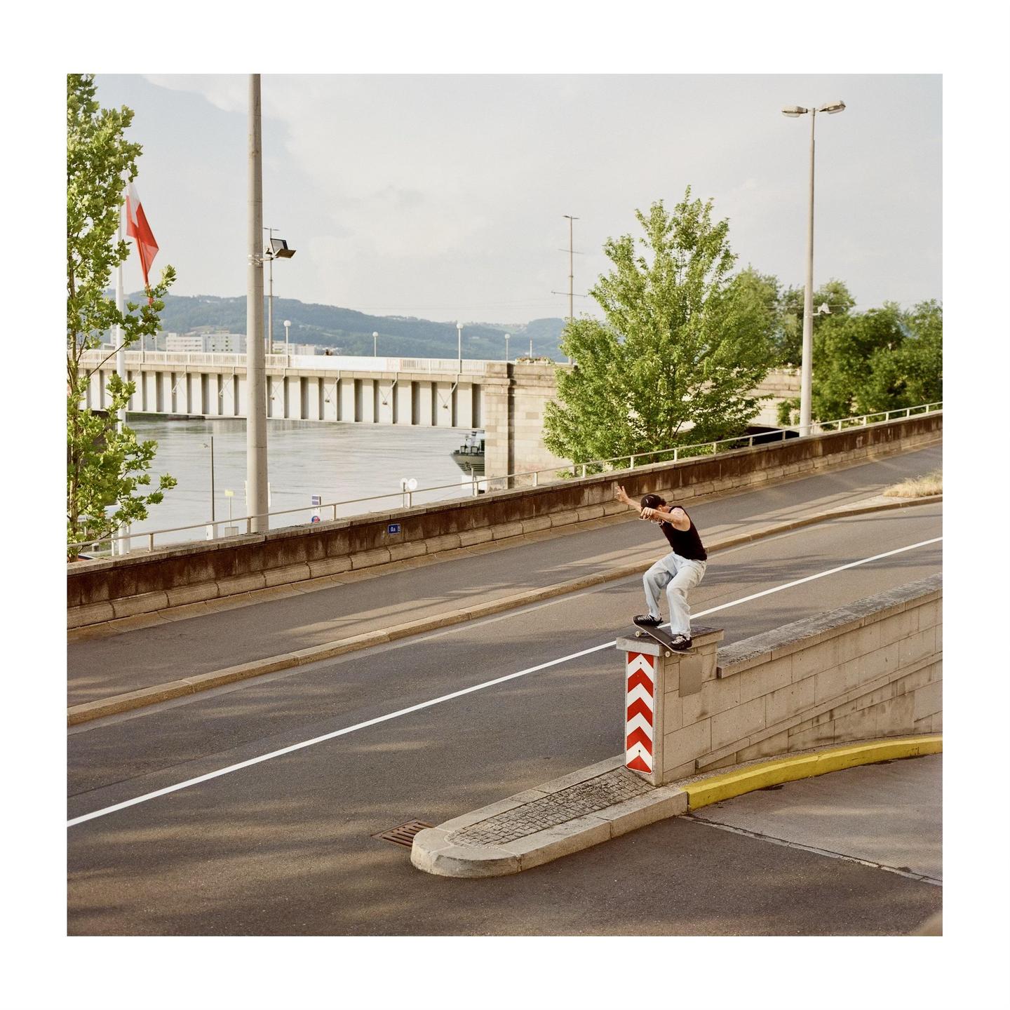 A man on a skateboard grinds a concrete ledge next to a road with a river and bridge in the background.