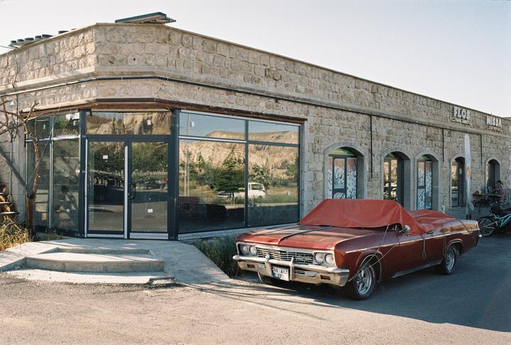 A red car with a cover over its roof is parked in front of a stone building with large windows reflecting a mountainous landscape.