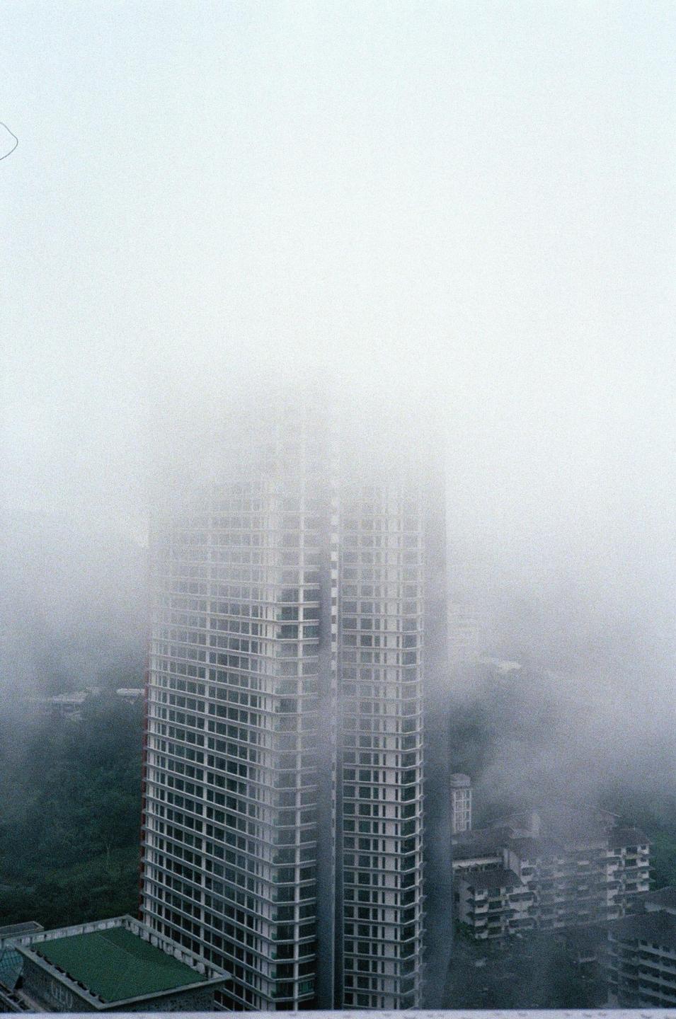 A tall building is partially obscured by fog, with other buildings and trees visible below.