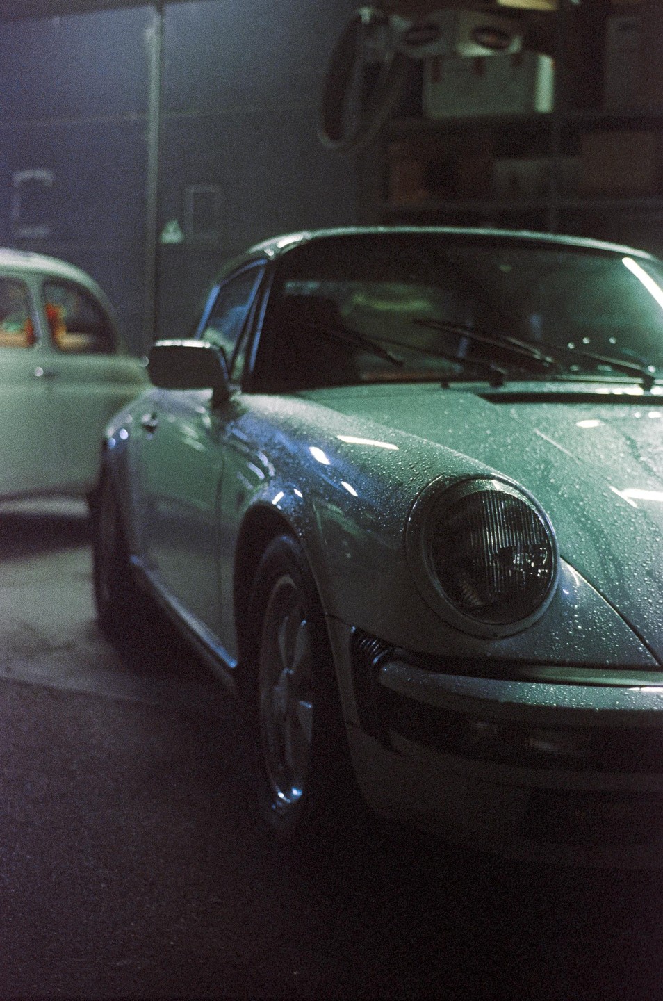 A light-colored vintage sports car covered in water droplets is parked in a dimly lit garage next to another car.