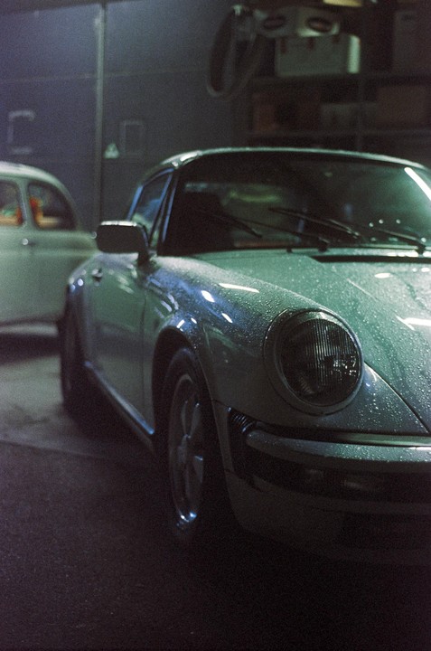 A light-colored vintage sports car covered in water droplets is parked in a dimly lit garage next to another car.
