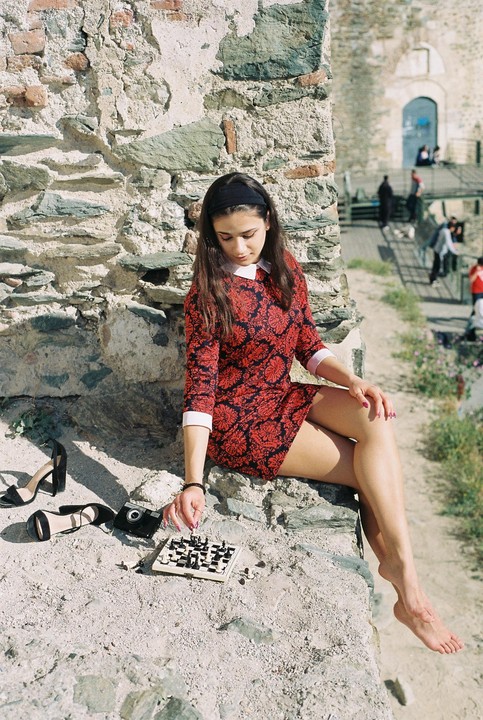 A woman in a red dress sits barefoot on a stone wall next to a miniature chessboard, with her shoes and a camera nearby.