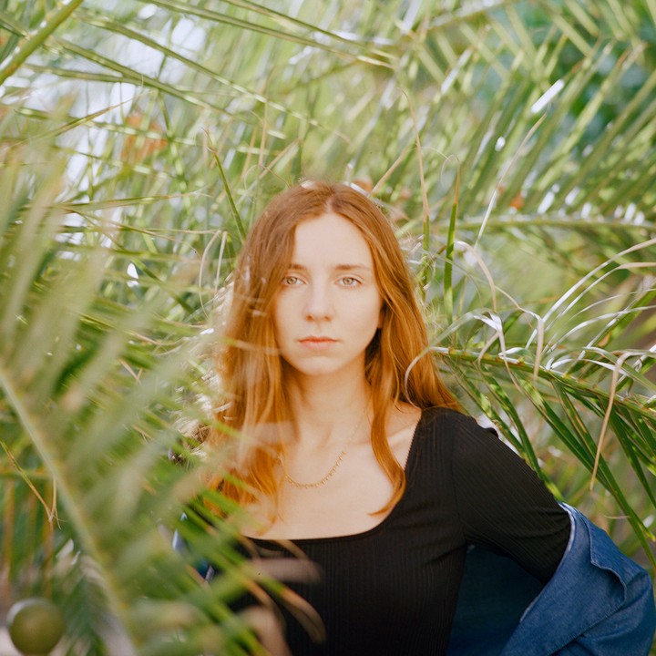 A woman with long hair looks directly at the viewer, surrounded by palm fronds.