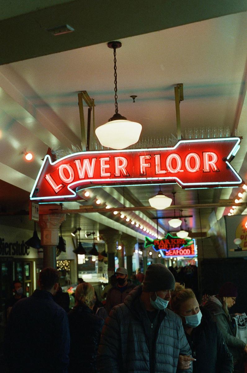A red neon sign points to the lower floor of a market where people are walking.