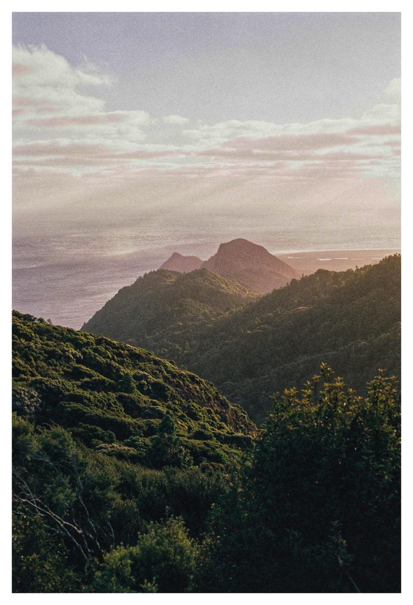 A mountain ridge covered in green foliage overlooks the ocean under a cloudy sky.