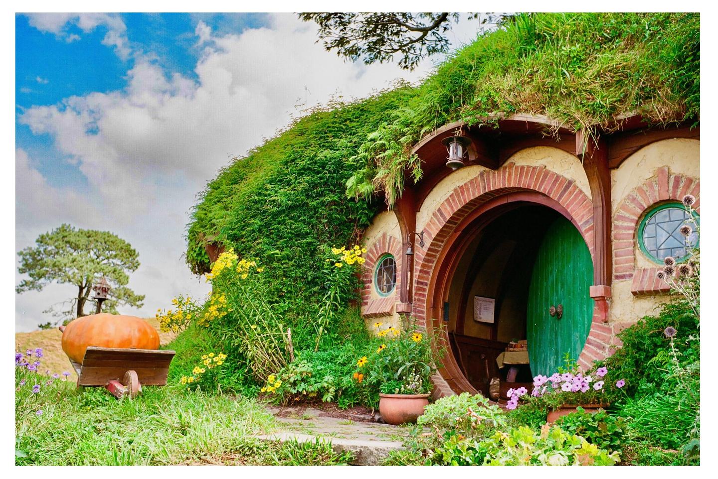 A hobbit hole with a round green door is nestled into a grassy hill, surrounded by flowers and a large pumpkin on a cart.