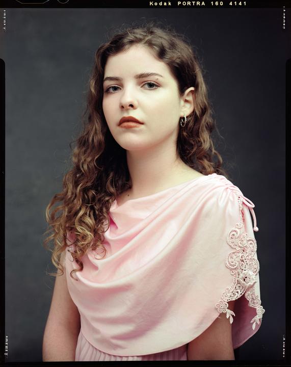 A young woman with curly hair wears a pink dress with lace details.