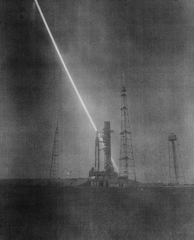 A long exposure image shows a rocket on a launchpad with a bright streak of light across the sky.