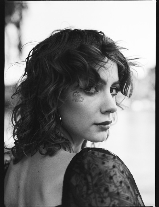 A woman with curly hair and a tattoo on her cheek looks over her shoulder.