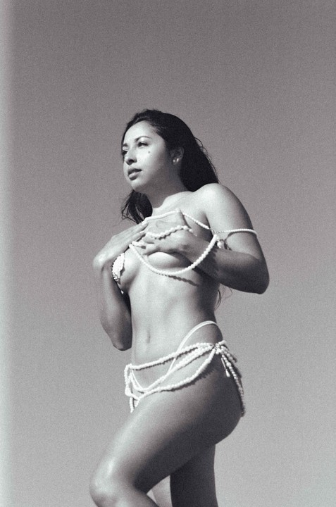 A woman poses topless, adorned with strands of pearls covering her chest and hips.