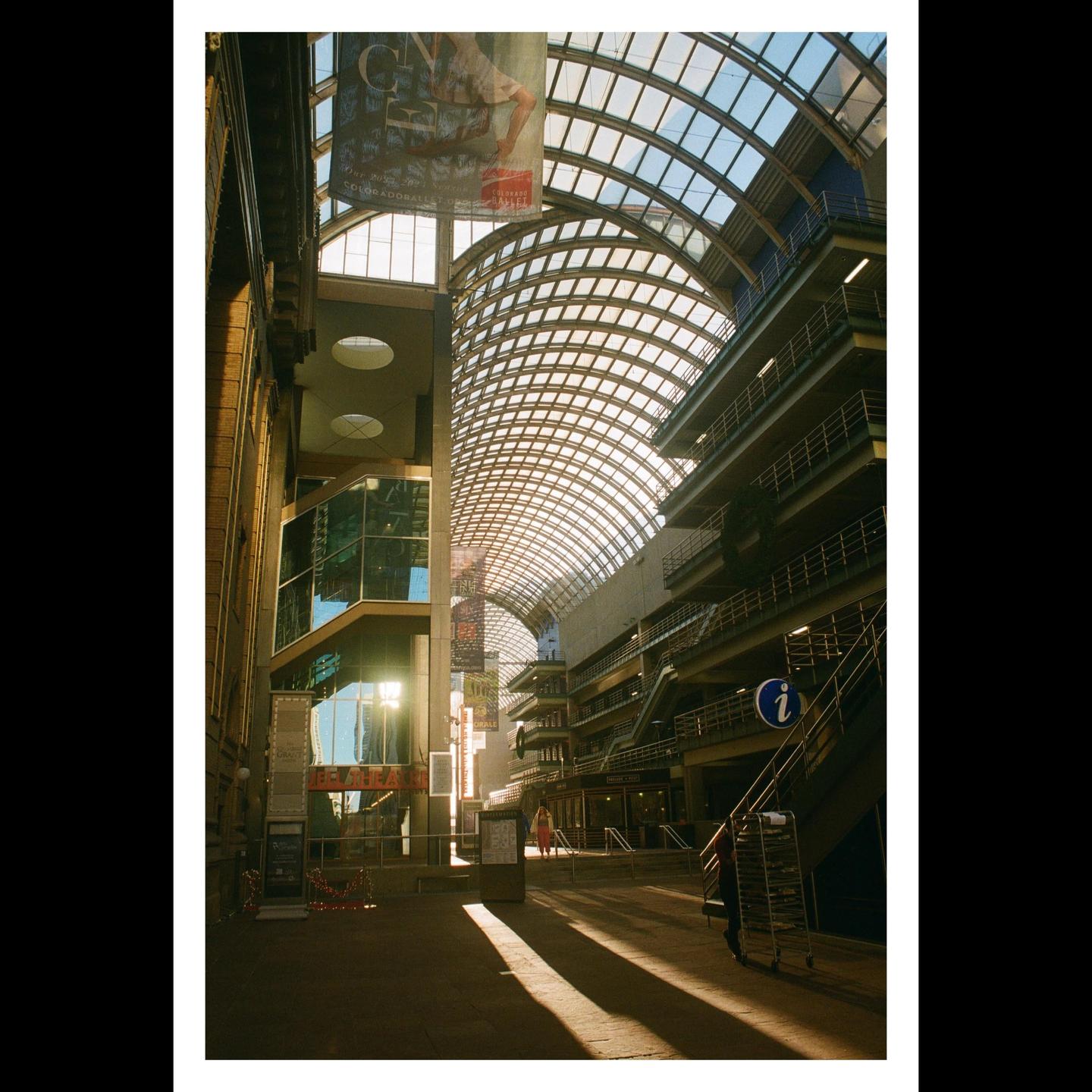 Sunlight streams through the glass arched roof of a large indoor space with multiple levels of balconies and a person walking in the distance.