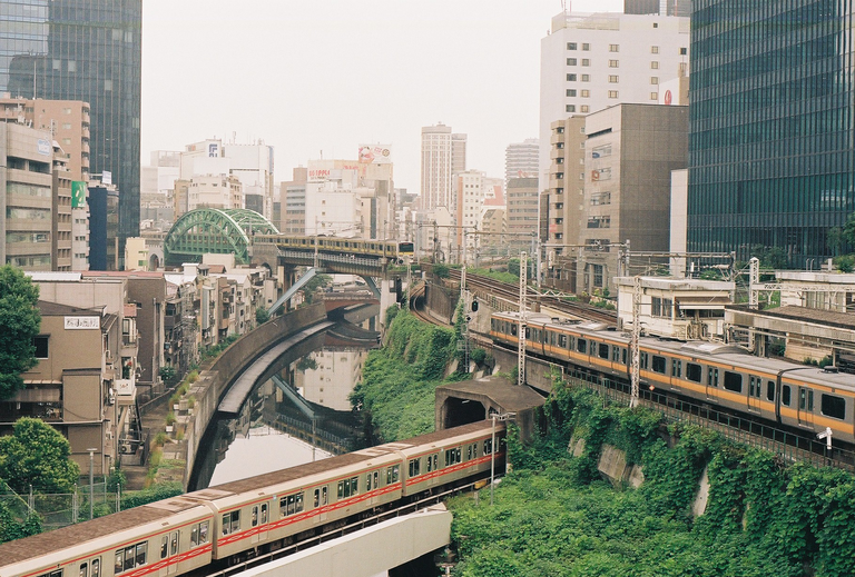 Three trains travel on tracks through a city with a river and buildings.