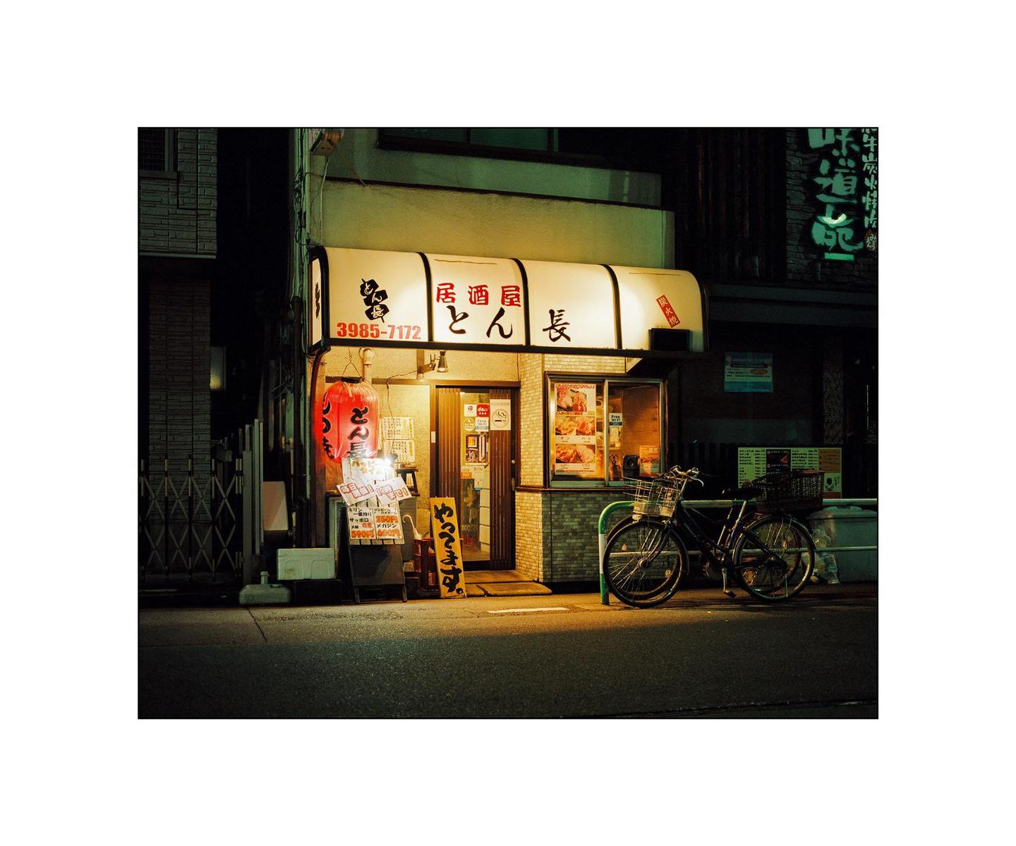 A bicycle is parked in front of a brightly lit Japanese restaurant at night.