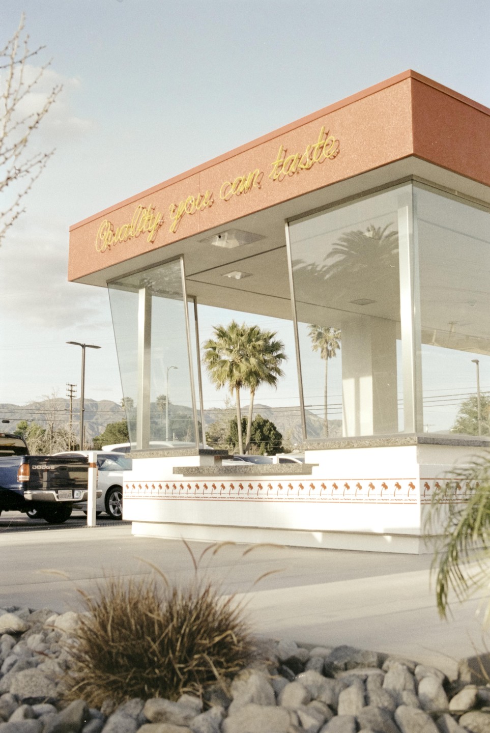 A building with a red roof and large windows reflects palm trees, with a truck and mountains in the background.