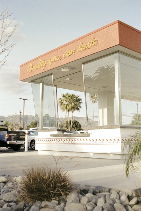 A building with a red roof and large windows reflects palm trees, with a truck and mountains in the background.