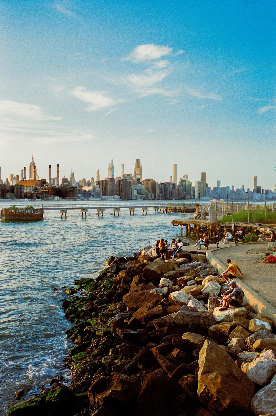 People sit on rocks and a path next to a body of water with a city skyline in the background.
