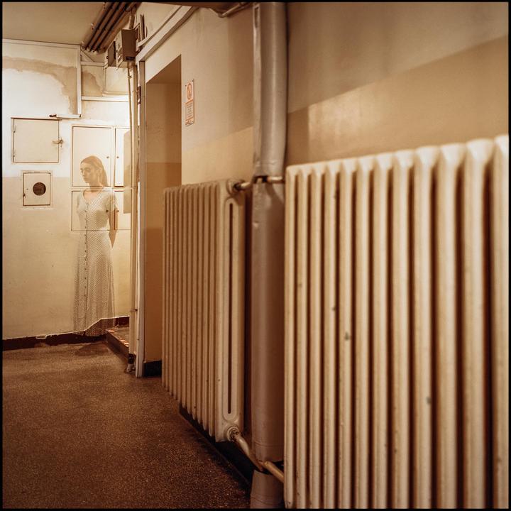 A woman in a long dress is visible through a doorway, with radiators lining the wall in the foreground.