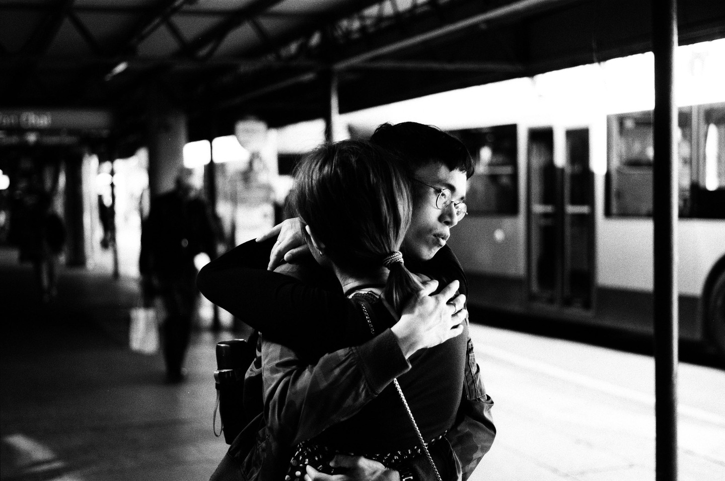 A couple embraces on a train platform with a train visible in the background.