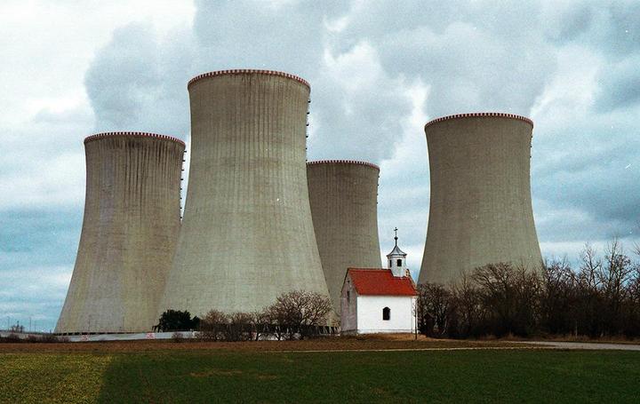Four cooling towers with smoke rising from them stand behind a small white chapel with a red roof.