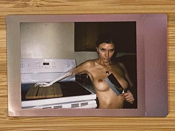 A topless woman holds a cleaver while a skeleton arm rests on a cutting board on a stove.