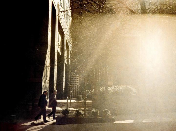 Two men walk past a building with strong shadows and bright sunlight.
