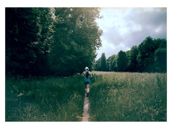 A woman runs on a narrow trail through a field of tall grass with trees on either side under a cloudy sky.