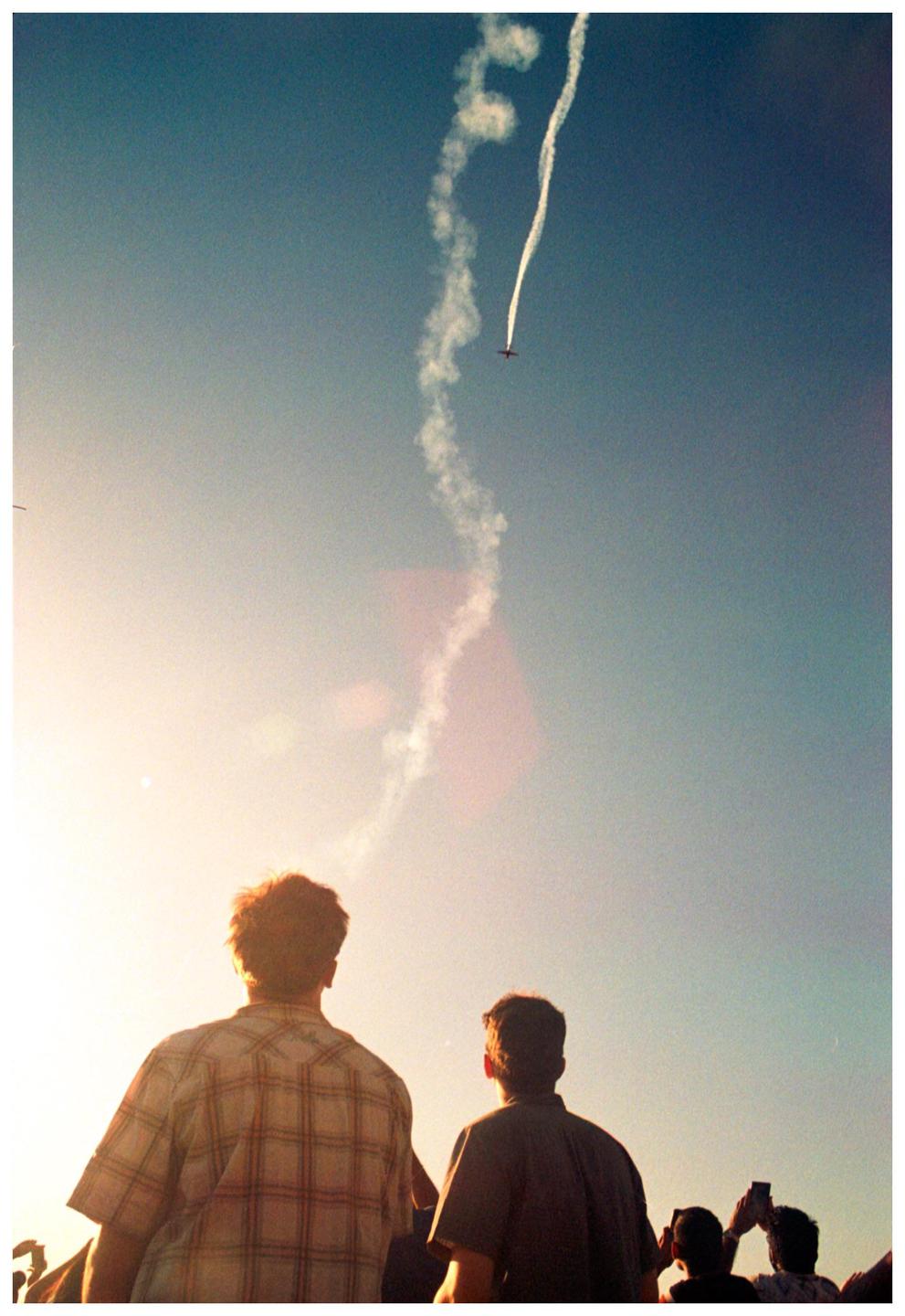 A crowd of people watches an airplane performing aerobatics with a smoke trail against a bright sky.