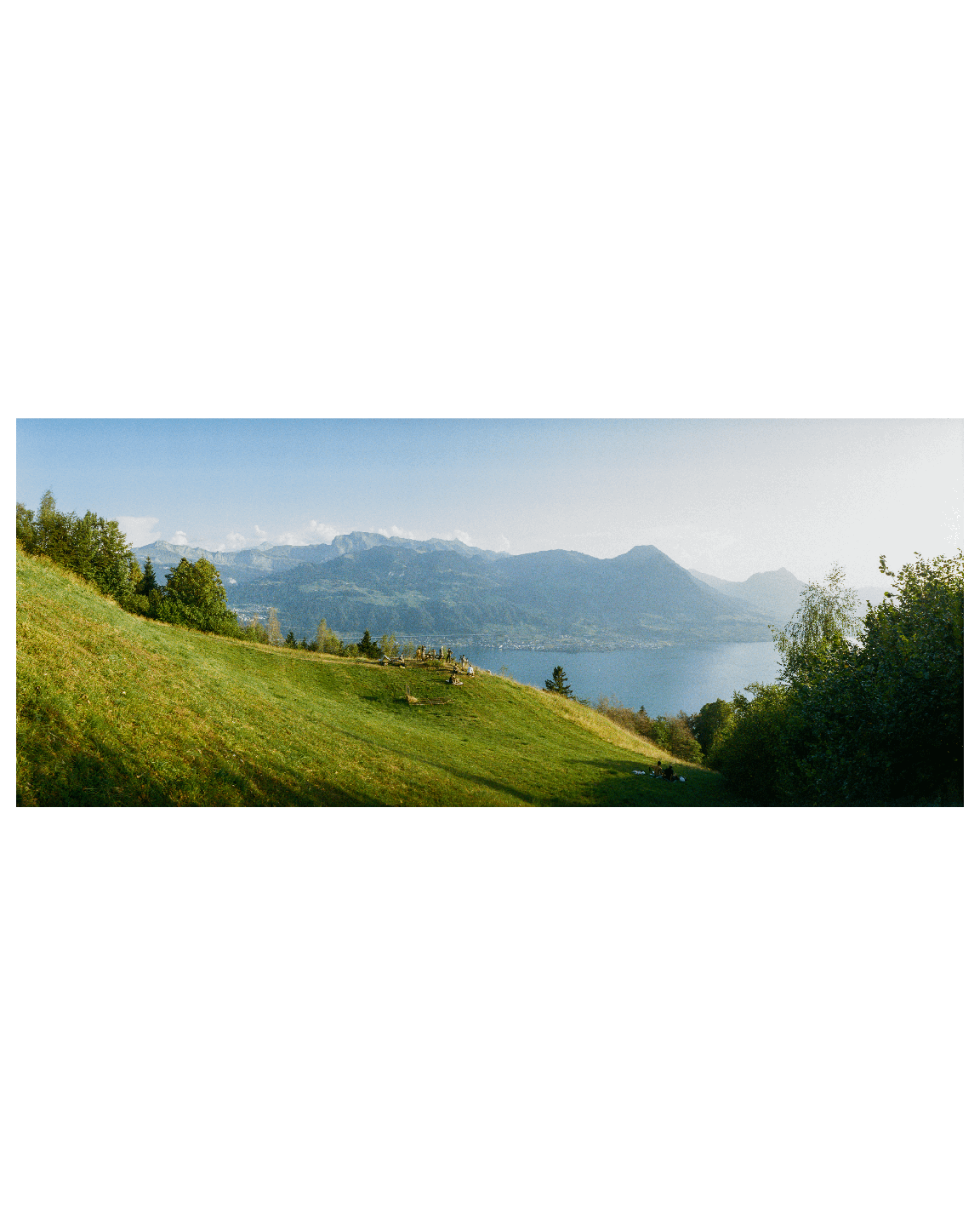 A panoramic landscape shows a grassy hillside overlooking a lake and mountains under a hazy sky.