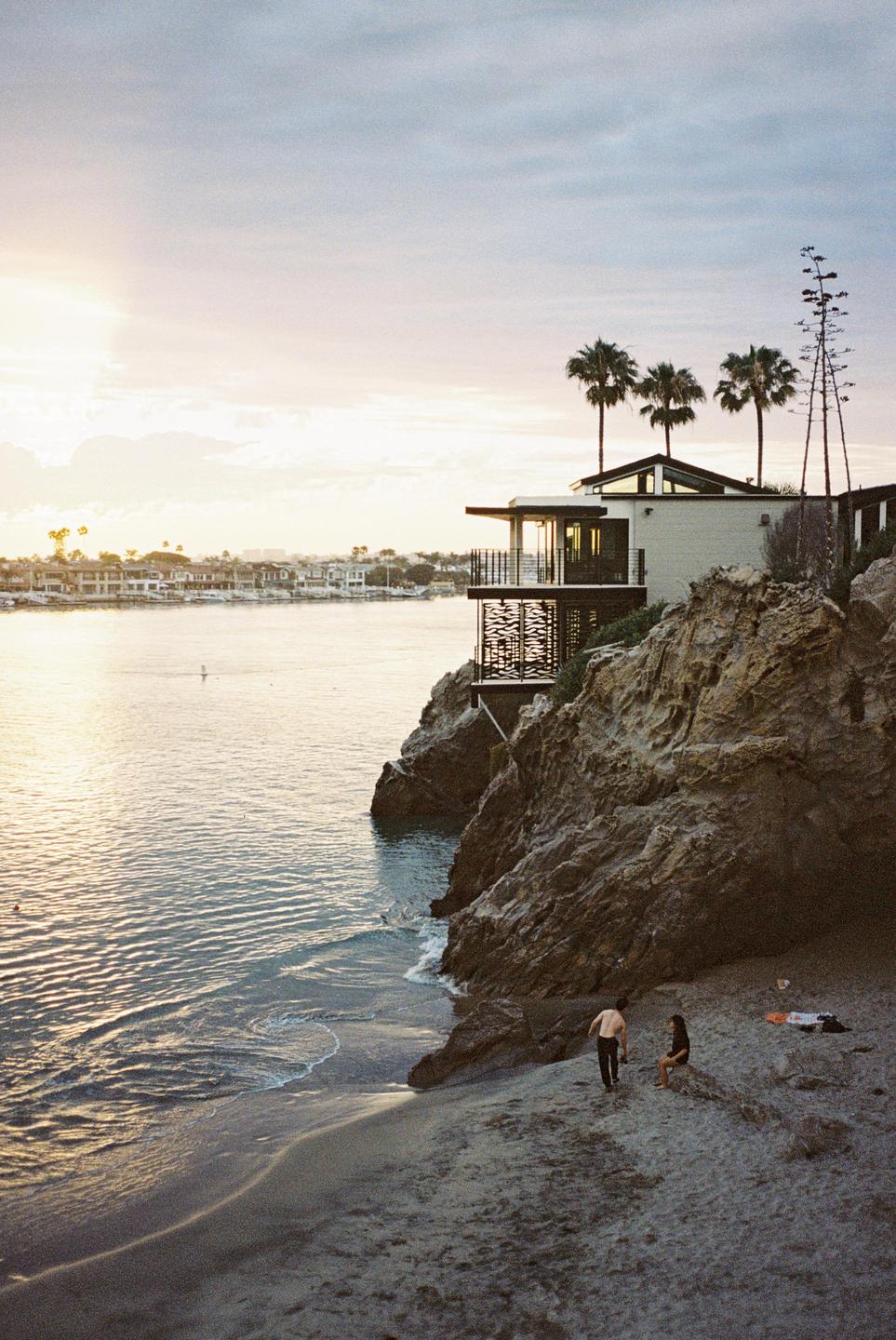 A modern house with palm trees sits on a rocky cliff overlooking a bay with a sandy beach where a couple is relaxing.