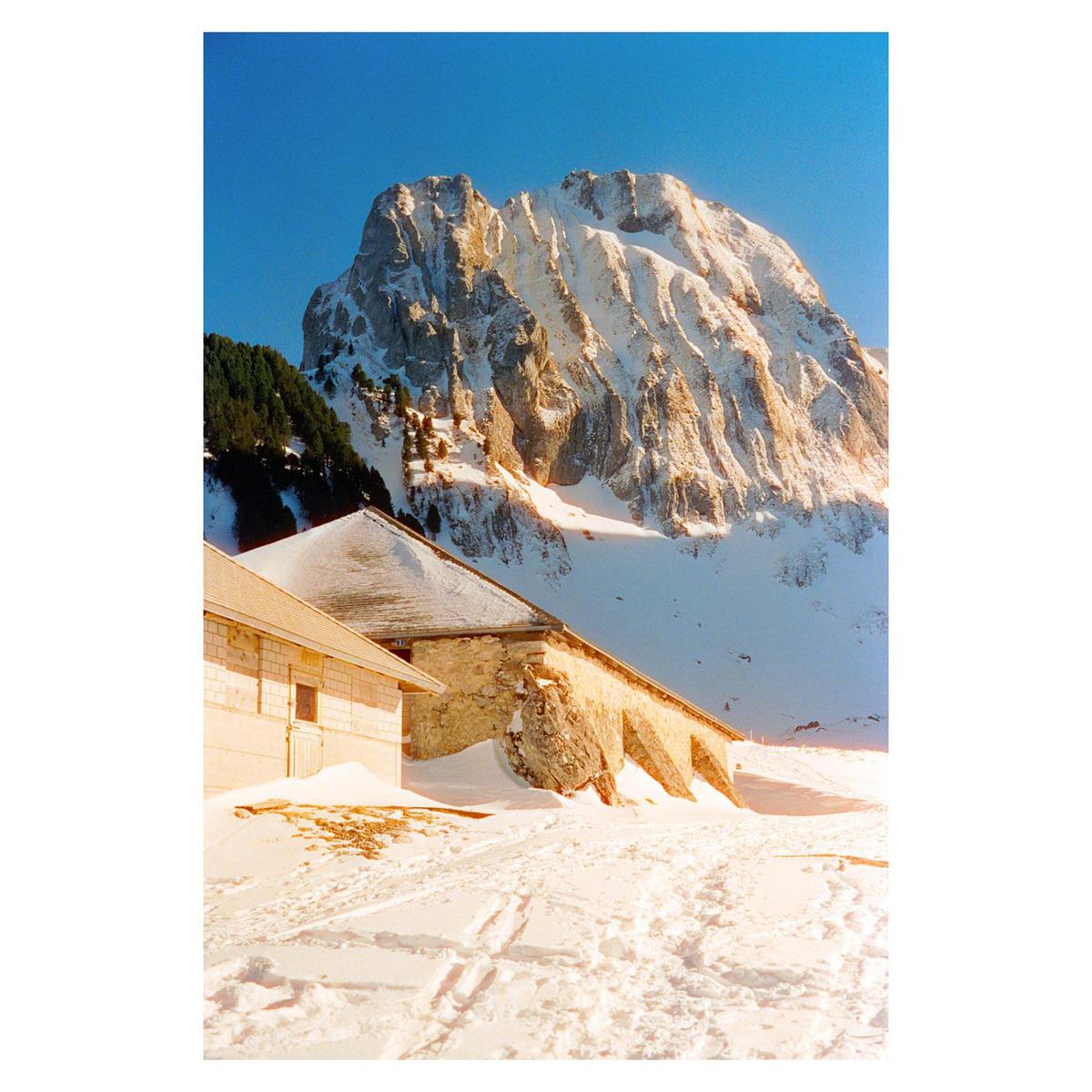 A snow-covered mountain rises behind a building with a snow-covered roof and a stone wall.