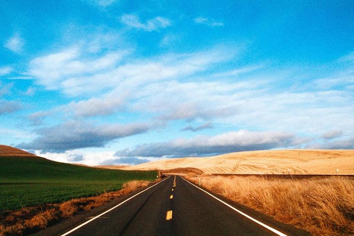 A long road stretches through rolling hills with green and golden fields under a blue sky with clouds.
