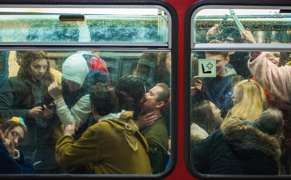 A couple kisses in a crowded metro car seen through a rain-streaked window.
