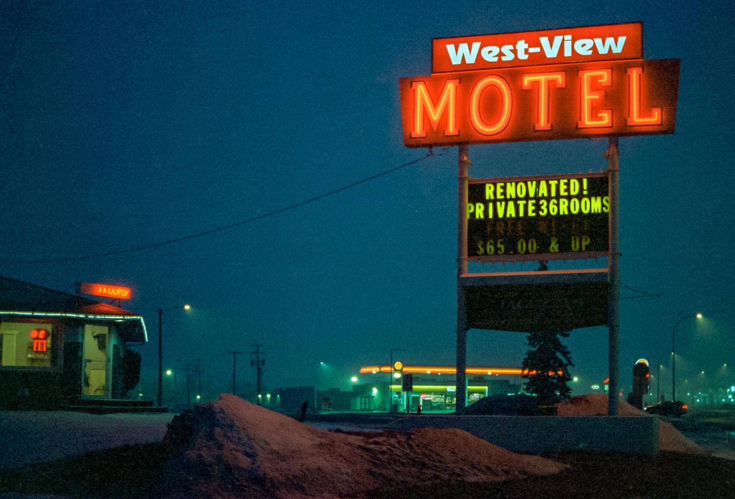 A neon motel sign stands in front of a building with a "vacancy" sign, with snow on the ground and a gas station in the background.