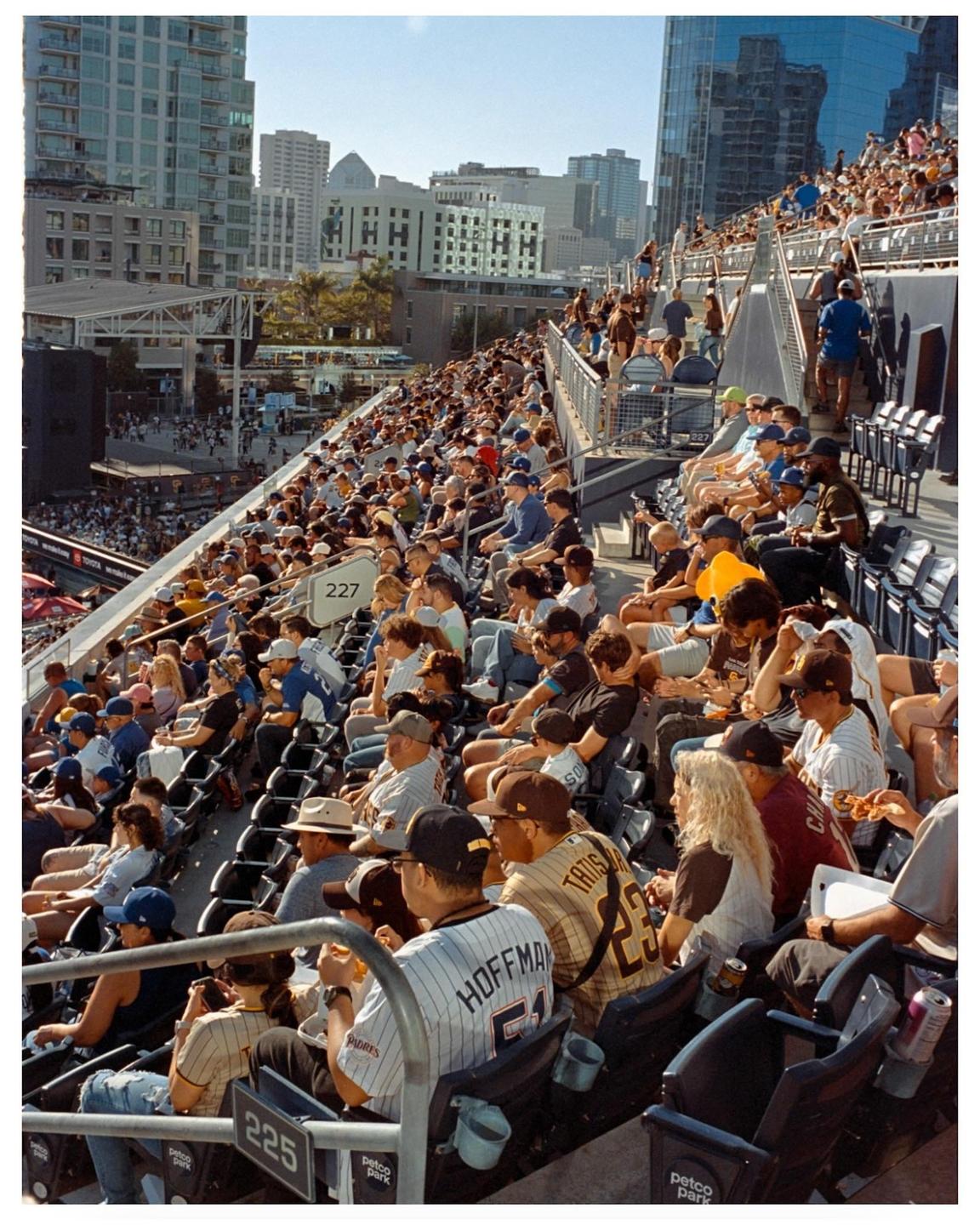 A large crowd of people are seated in a stadium with city buildings in the background.