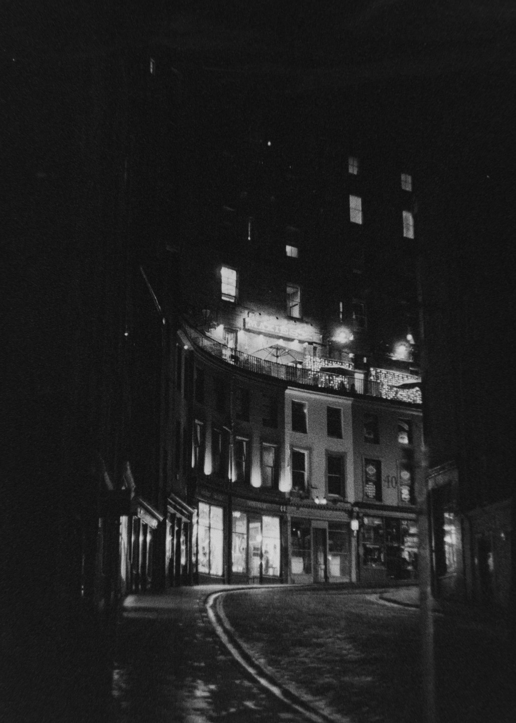 A street curves past buildings with illuminated windows and a balcony at night.