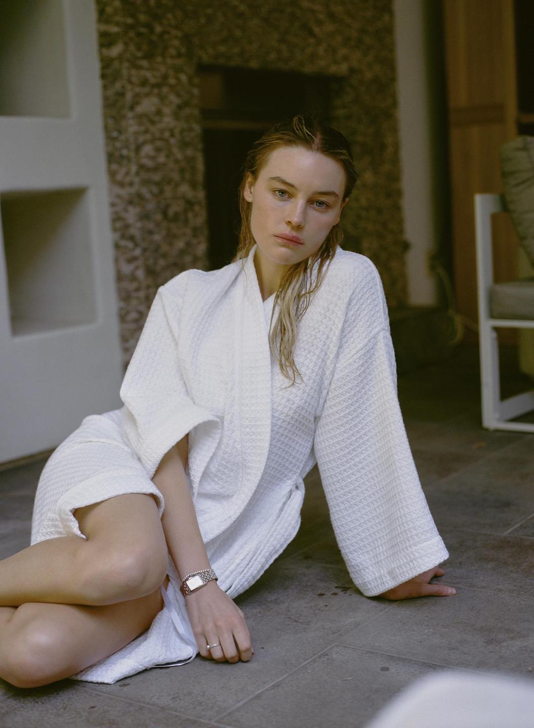 A woman in a white robe sits on a tiled floor looking directly at the viewer.