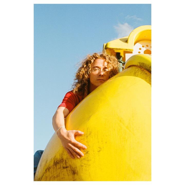 A man with curly hair and glasses is embracing a large yellow object under a clear blue sky.