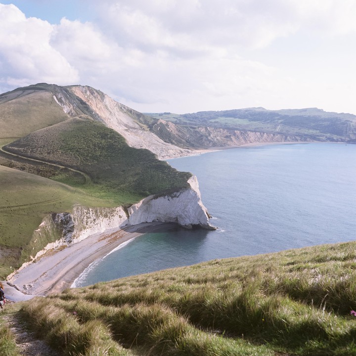 A person walks on a grassy hill overlooking a bay with white cliffs and a beach.