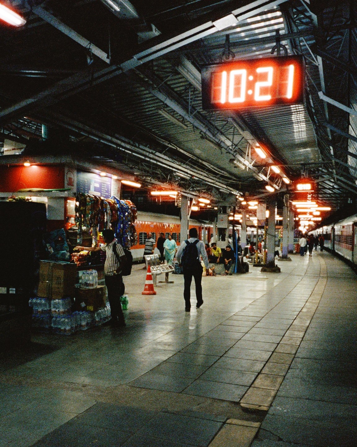 A digital clock displaying 10:21 hangs above a busy train platform at night.