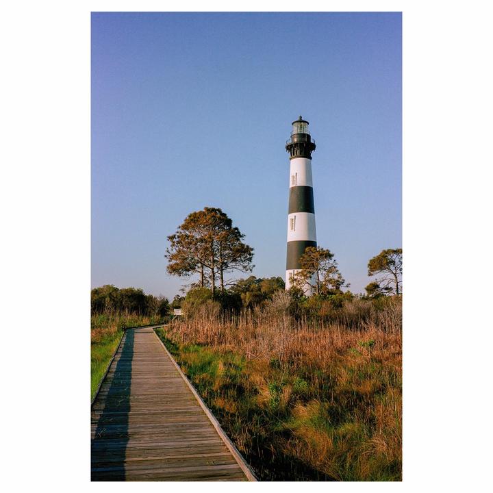 A black and white striped lighthouse stands tall against a clear blue sky, next to a wooden boardwalk leading through a grassy landscape.