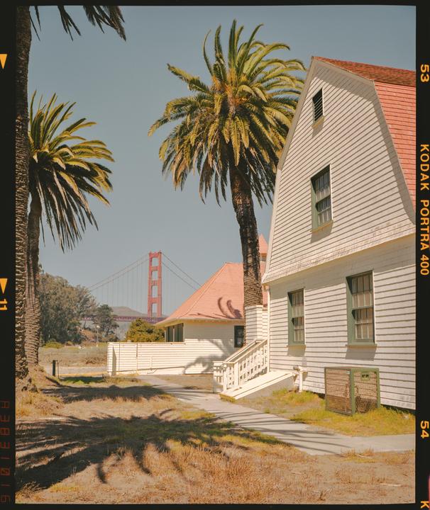 A white house with a red roof stands next to palm trees, with the Golden Gate Bridge visible in the background.