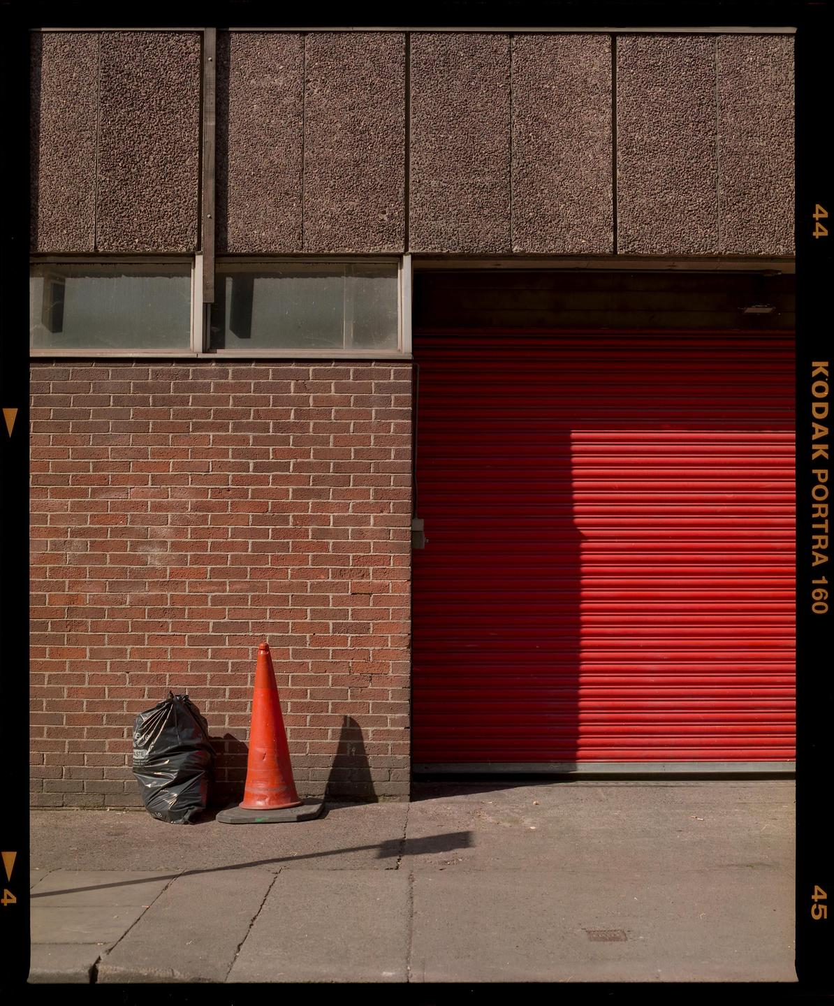 A red traffic cone and a black garbage bag sit on a sidewalk next to a brick wall and a red garage door.