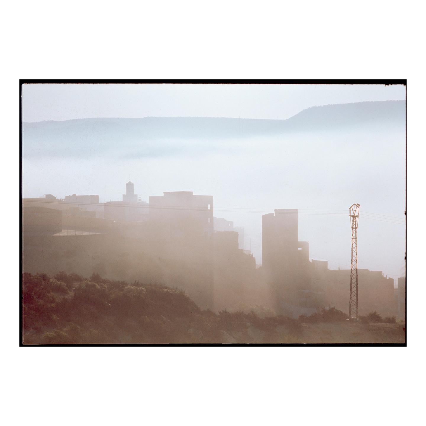 A town with a tall tower is shrouded in fog with mountains in the background.