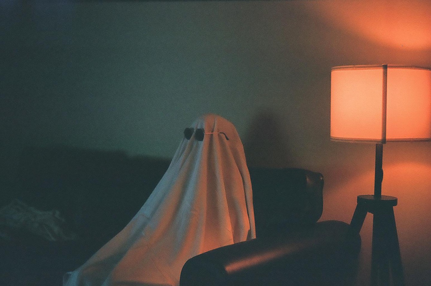 A person dressed as a ghost with heart-shaped sunglasses sits on a couch next to a lamp.