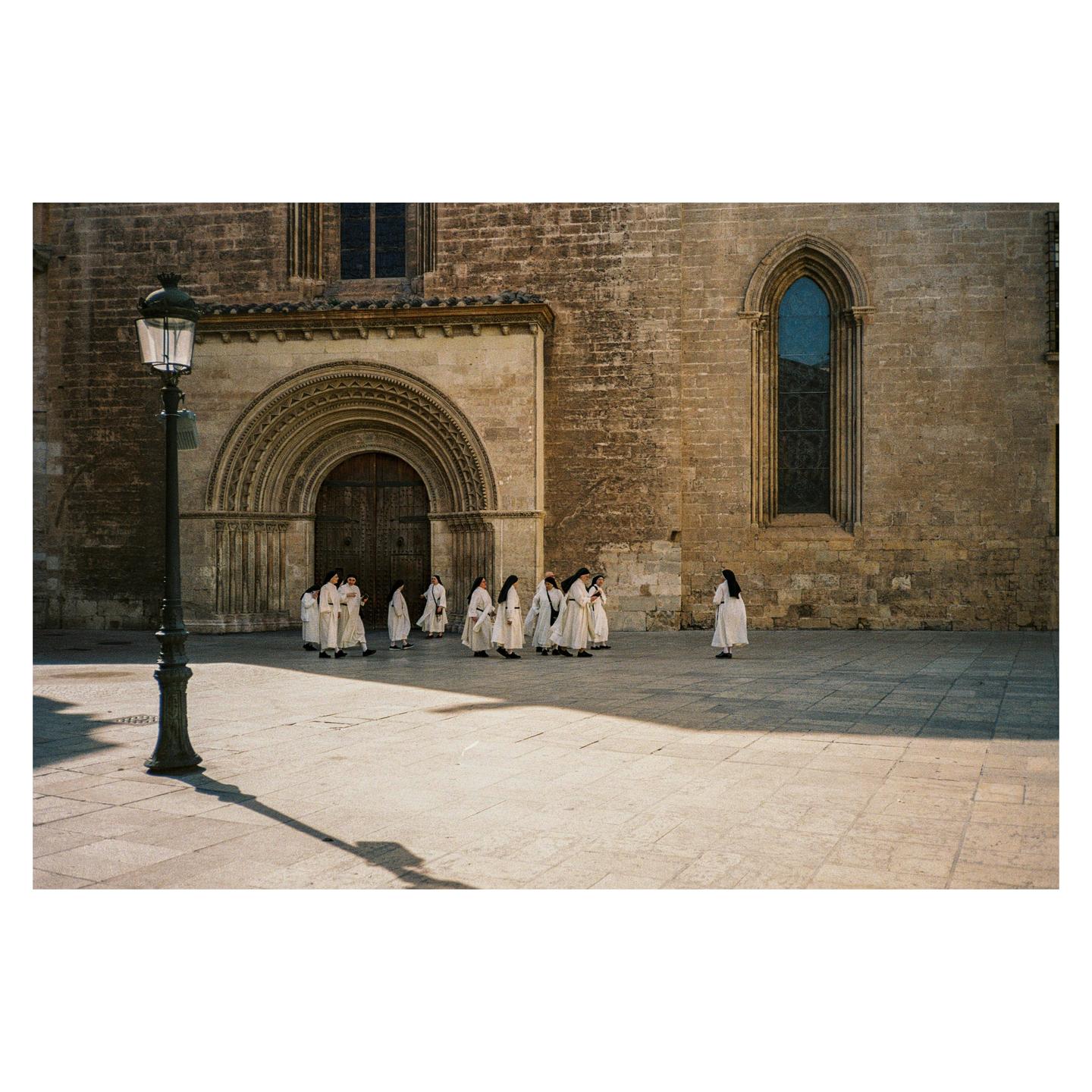 A group of nuns in white habits walk across a stone plaza in front of a large stone building with an arched doorway and tall window.